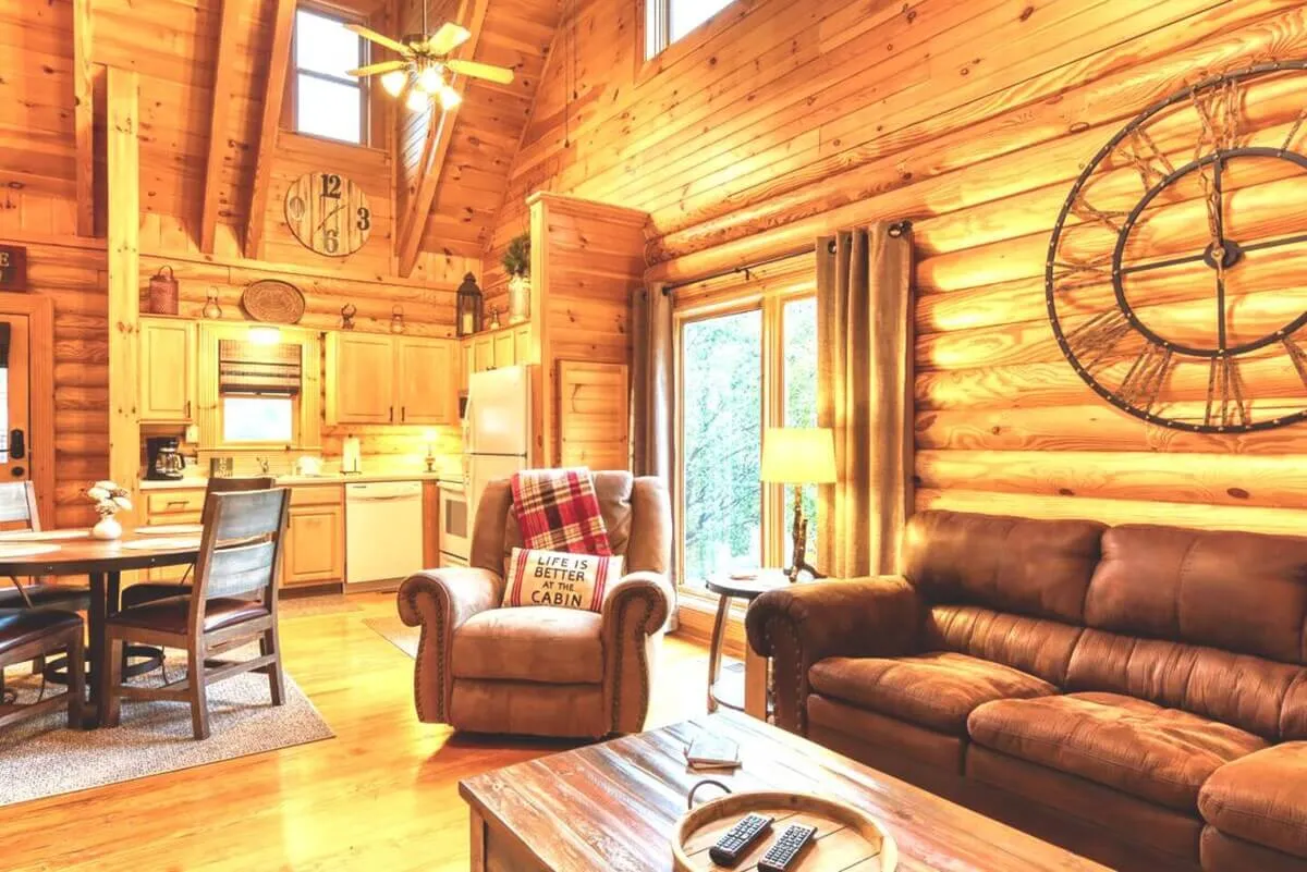 A cozy, open-plan space with wooden walls, a comfortable seating area, a large clock, a welcoming dining table, and expansive windows that fill the room with natural light. The setting feels ideal for family gatherings or evenings spent relaxing together.