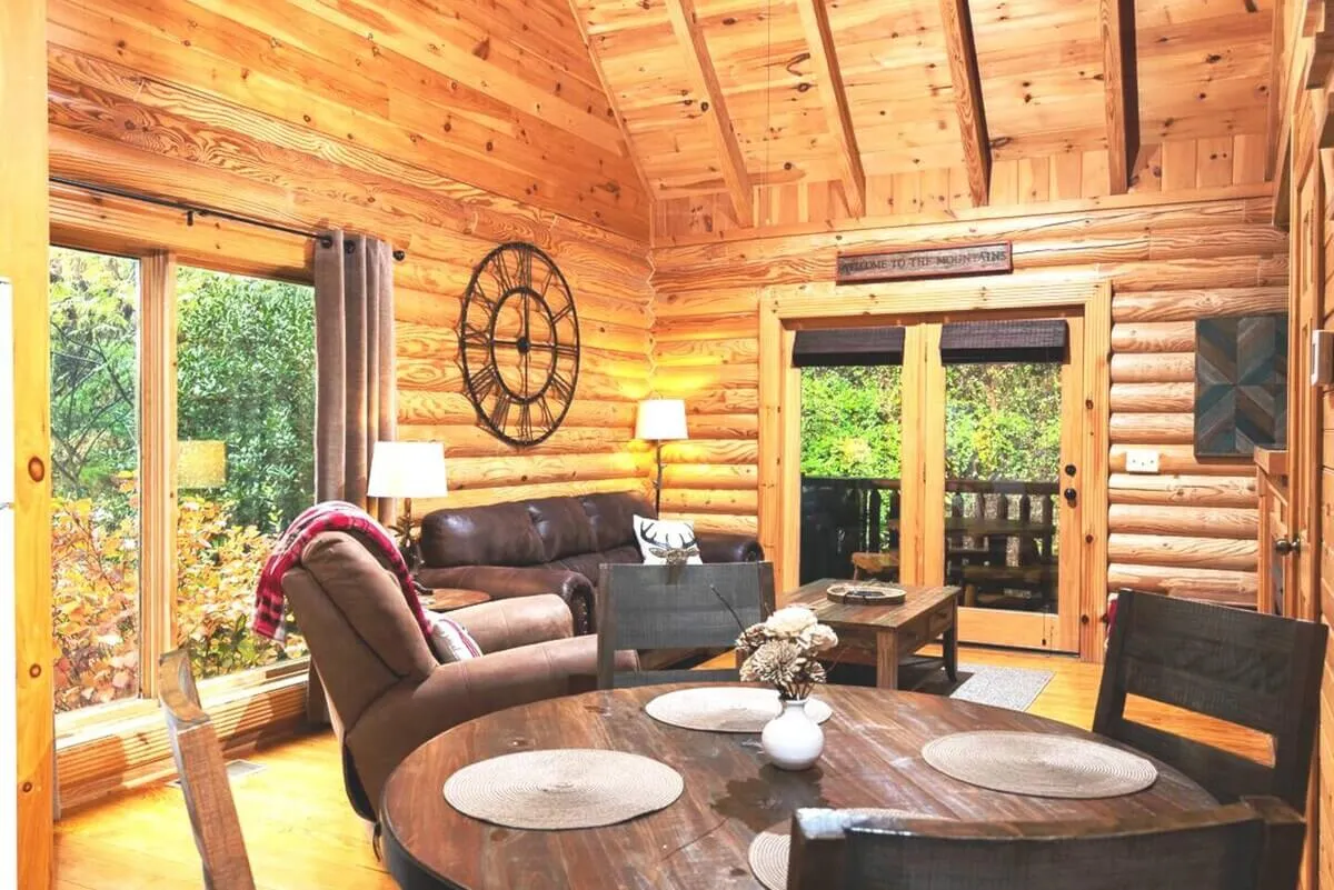 A warm, inviting living area featuring wooden log walls, a comfortable leather sofa, large windows looking out onto nature, a round dining table perfect for group meals, and a decorative wall clock. The room is bathed in natural light and designed for social gatherings and relaxation.