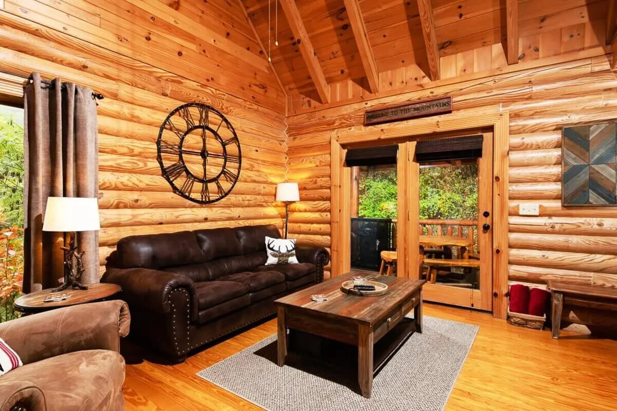 A brown leather sofa anchors this cozy living space, complemented by a wooden coffee table, a wall clock, and open doors leading to the outdoors. The room is designed for relaxation, reading, and enjoying the warmth of the cabin with the possibility of taking in sunset views from the adjacent deck.