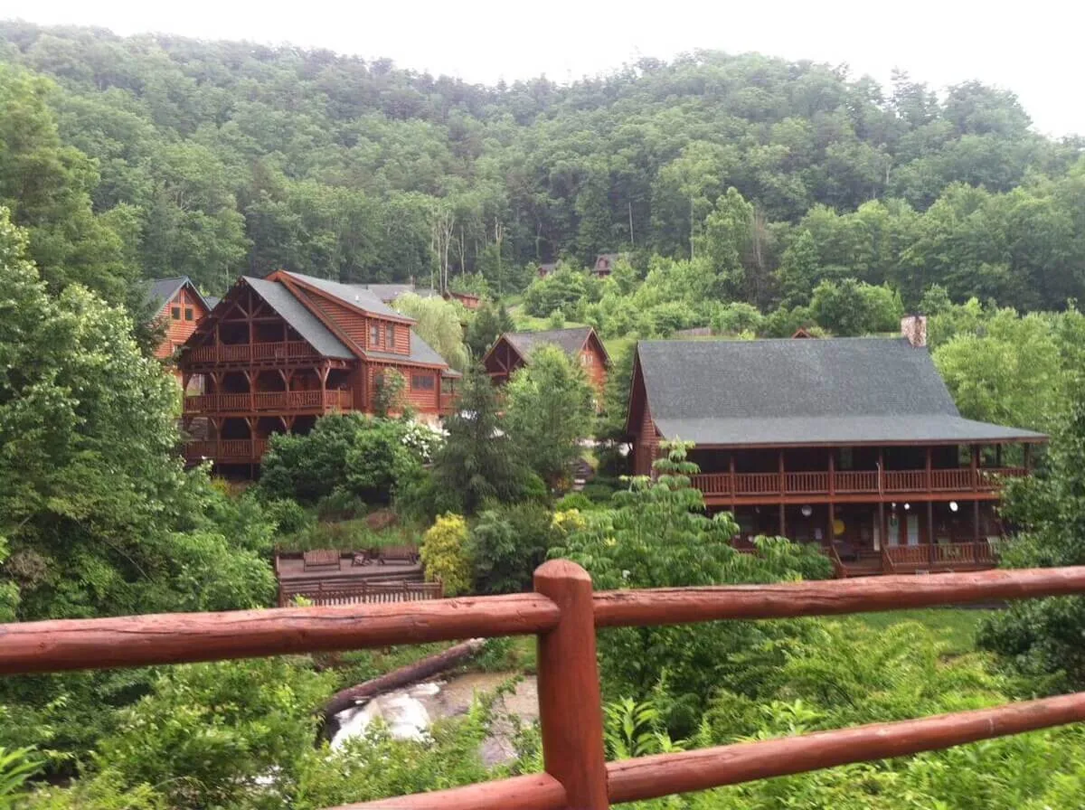 A charming row of log cabins surrounded by lush greenery, with a nearby stream and wooden railings that invite exploration. The cabins are set in a peaceful, wooded area, suggesting both adventure and relaxation.