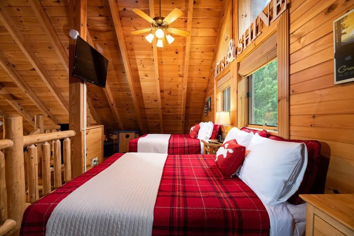 A warm and welcoming bedroom featuring rustic wooden beams, plaid bed covers, and nightstands with softly glowing lamps. Natural light filters through a window, illuminating the inviting textures and cozy decor. The space exudes comfort, with a harmonious blend of wood and textiles that evoke a mountain retreat feel.