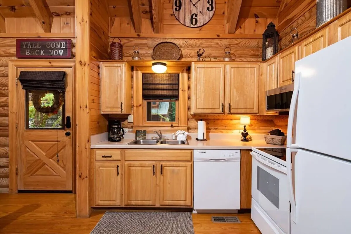 The kitchen features wooden cabinetry, up-to-date appliances, a decorative wall clock, and an entry door that hints at an open and accessible layout. The work surfaces are uncluttered, making the space feel organized and ready for culinary adventures or group gatherings.