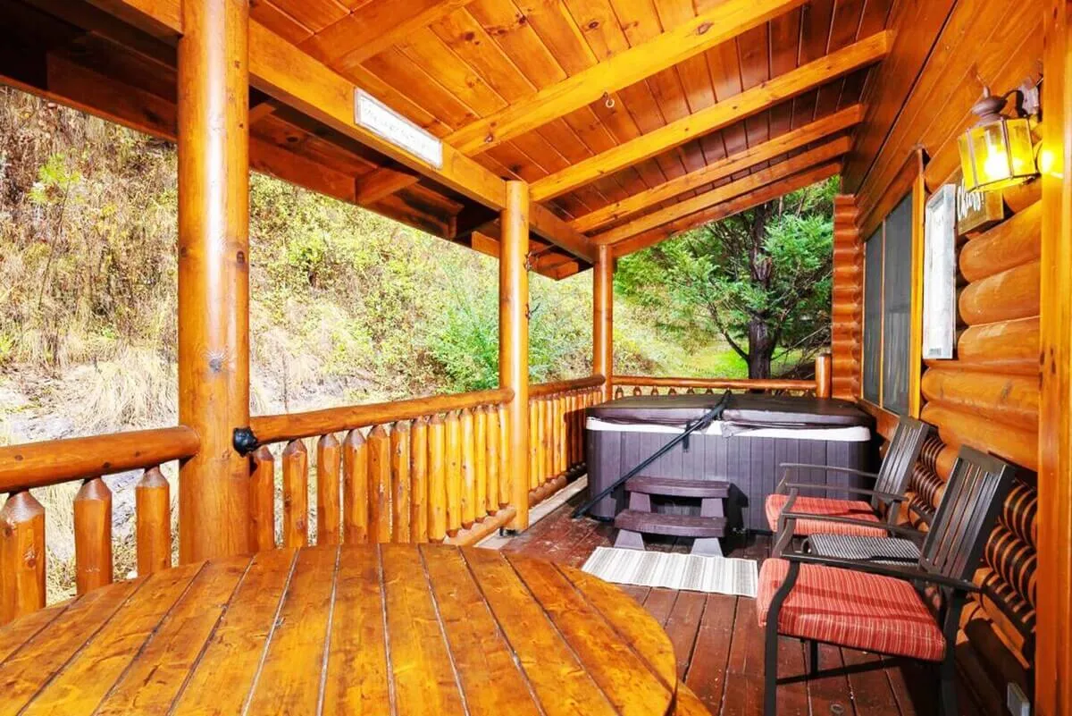 This image showcases a private wooden deck featuring a bubbling hot tub, an outdoor wooden table, and comfortable seating, all surrounded by lush green woods. The setting evokes a peaceful retreat, ideal for relaxing in the hot tub under the stars or enjoying meals outdoors while immersed in nature.