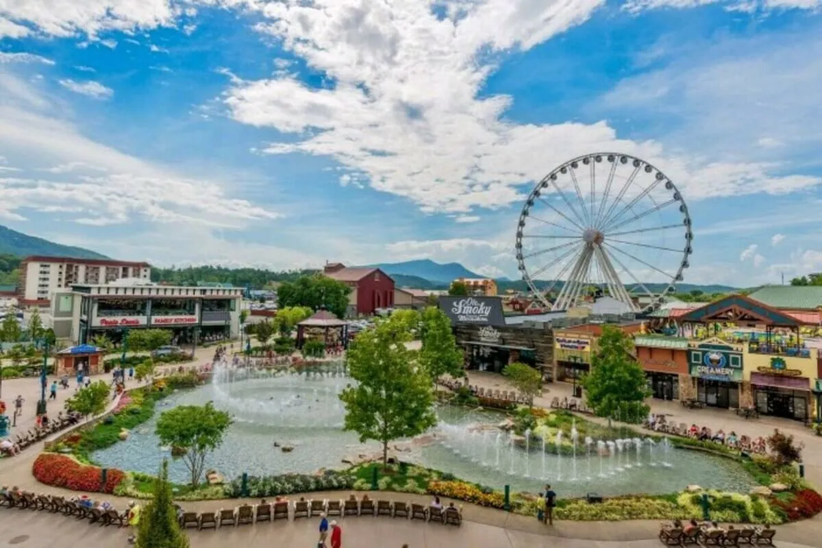A lively exterior scene featuring a large ferris wheel, a sparkling fountain, a pond, surrounding mountains, colorful flowers, and groups of people enjoying the attractions. The atmosphere is festive and inviting, echoing the bustling family fun at places like The Island and Dollywood.
