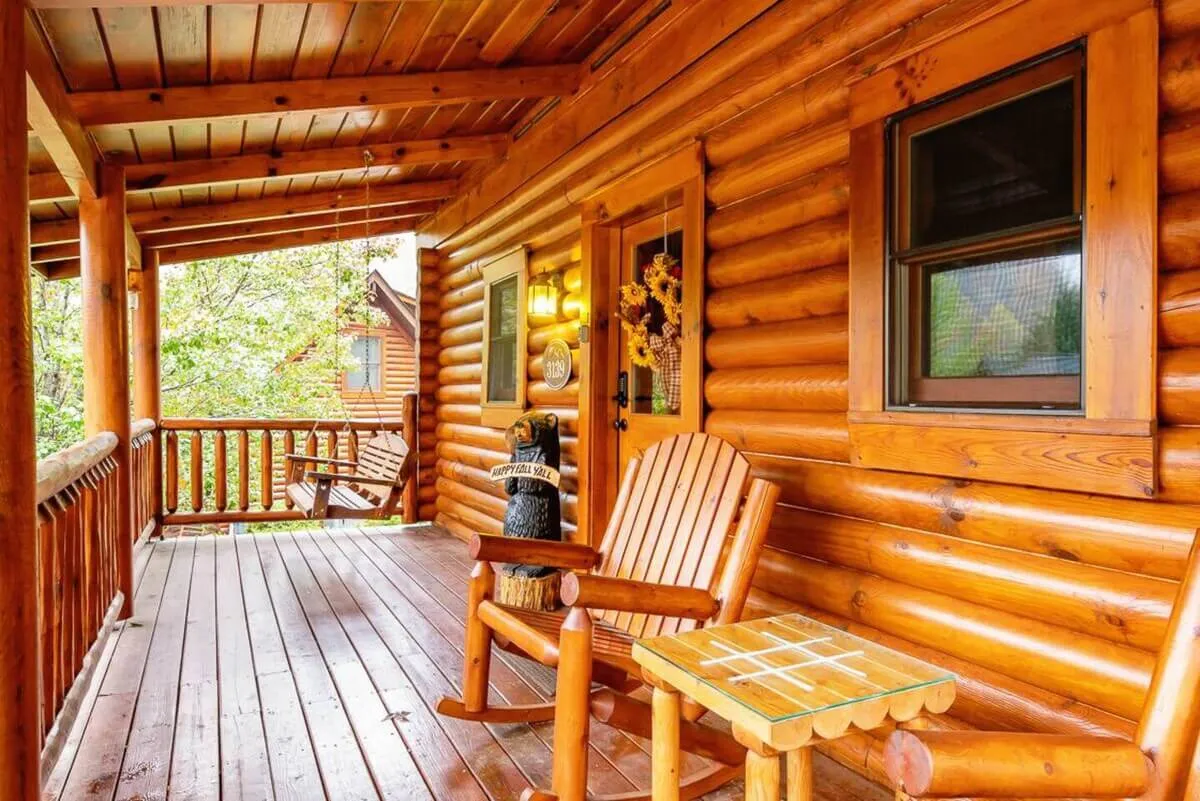 The image displays a log cabin deck with classic wooden rocking chairs and a small wooden table, adorned with decorative flowers. The cozy scene invites guests to sip coffee, read, or simply enjoy the sounds of nature, with the surrounding woods providing a calming backdrop.