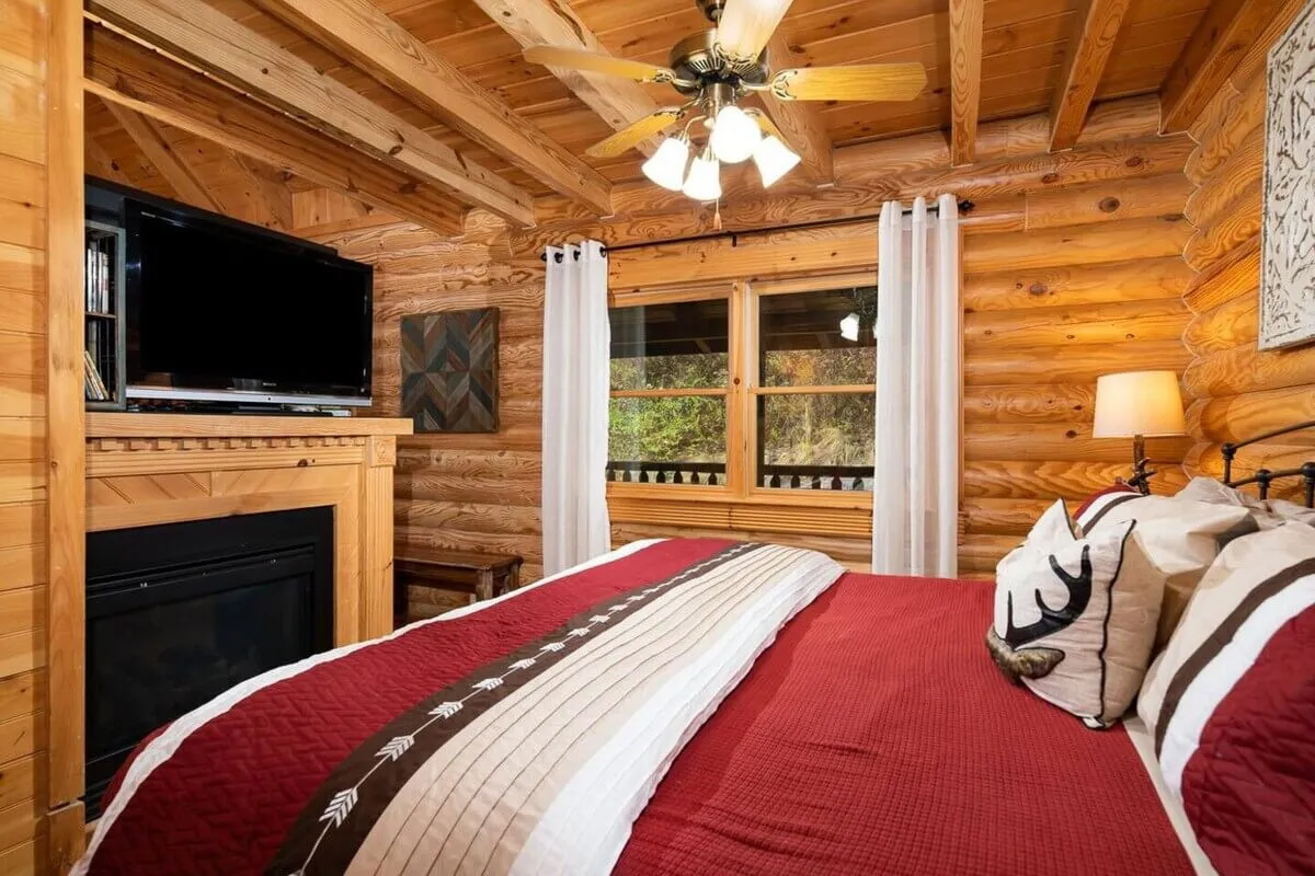 A bedroom with a red coverlet on the bed, wooden walls, a large window with curtains, and a fireplace. A television sits nearby, creating a multi-functional yet intimate retreat. The space is bathed in warm light, with a view through the window that hints at the surrounding landscape.